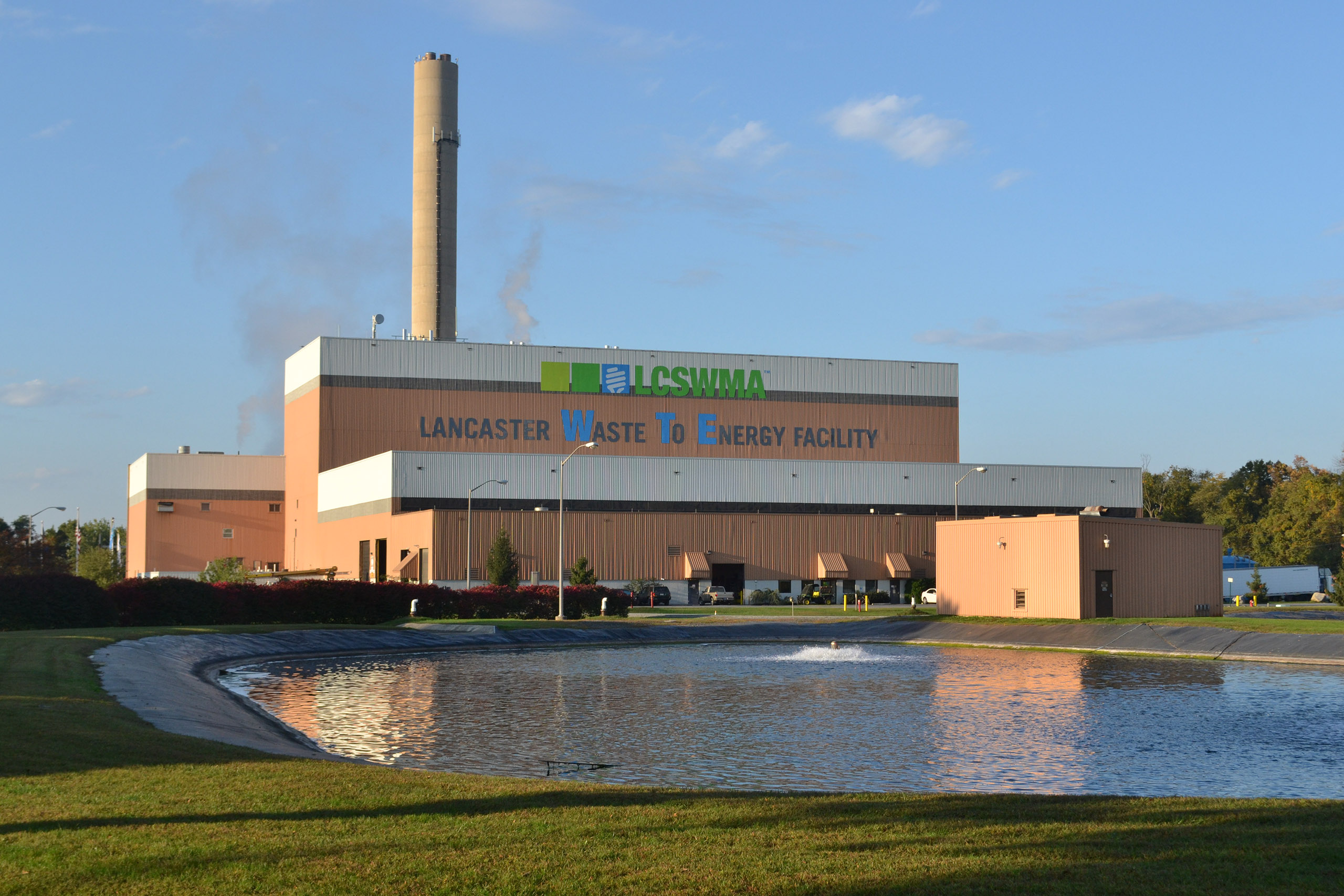 large lcswma building labeled Lancaster Waste to Energy Facility with a tall smokestack, situated beside a reflecting pond under a blue sky.