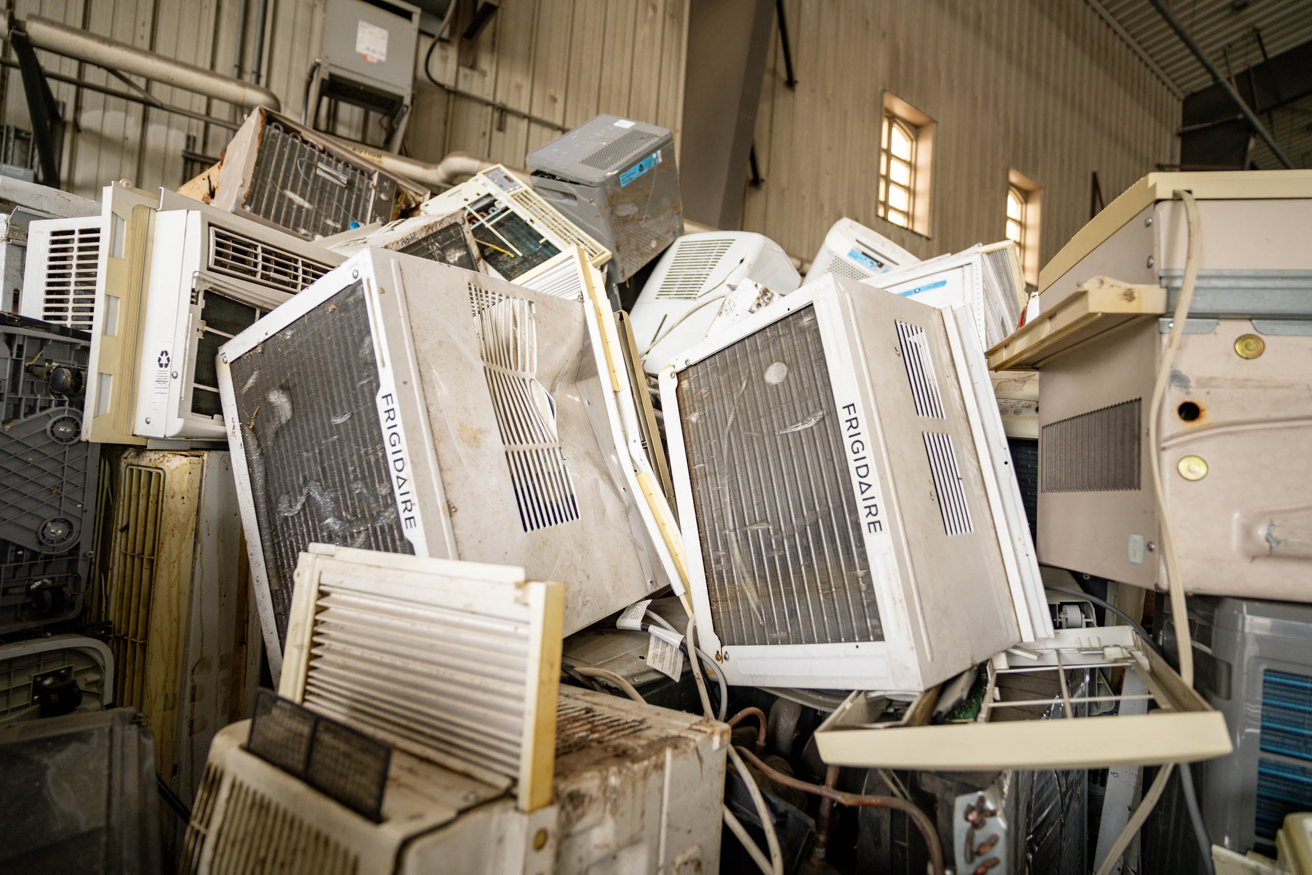 A large pile of old, discarded window air conditioning units stacked inside a warehouse awaits hauler pickup, with visible brand names and metal walls in the background.