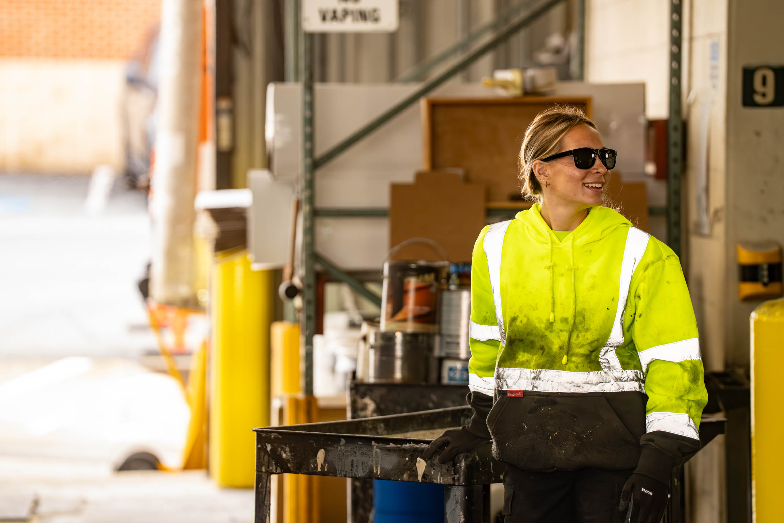 A woman wearing a bright yellow reflective jacket, black gloves, and sunglasses stands smiling next to a work table in an industrial setting.