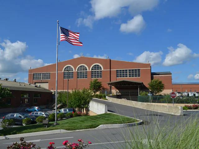 A large brick building with arched windows, an American flag flying in front, and several cars parked in a lot on a sunny day with blue sky and scattered clouds—explore more through Virtual Facility Tours of LCSWMA Facilities.