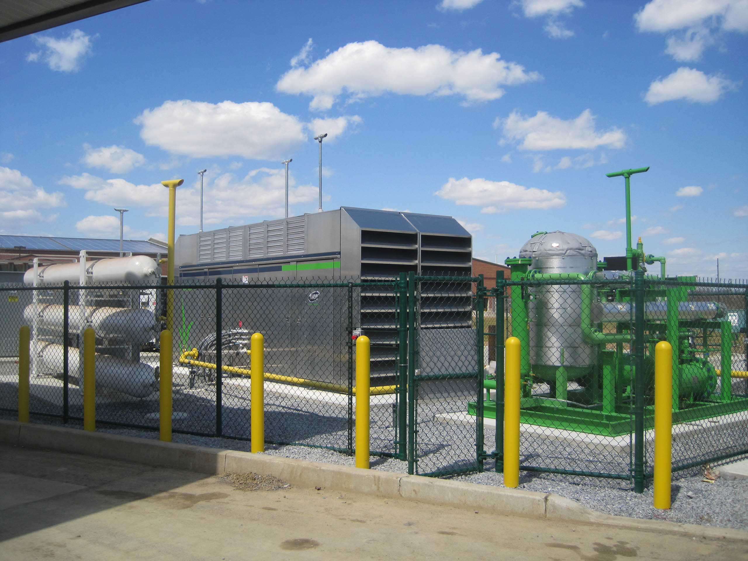 Industrial waste equipment, including large metal machinery, pipes, and tanks for Compressed Natural Gas (CNG for Businesses), sits behind a green chain-link fence with yellow posts outdoors under a blue sky with scattered clouds.