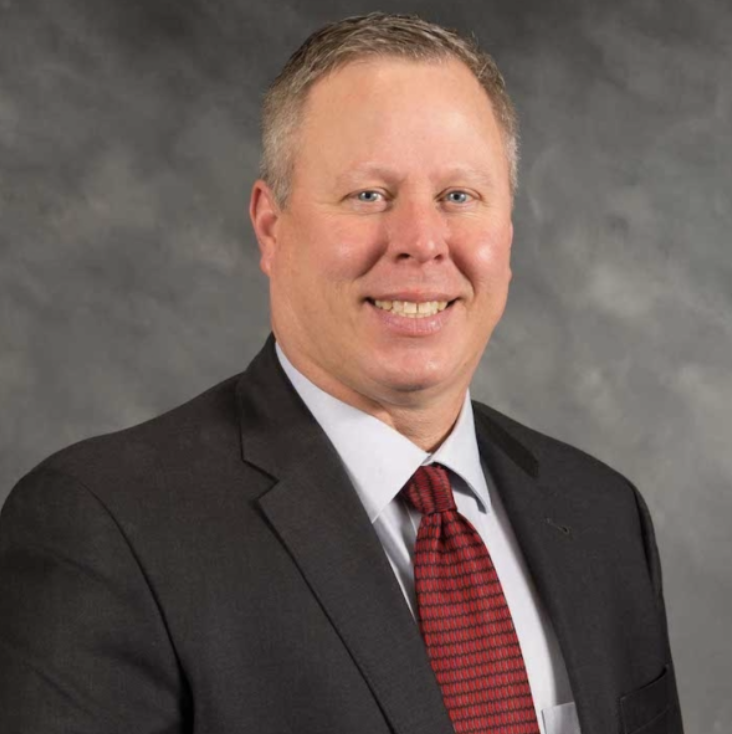 A man in a dark suit, light blue shirt, and red patterned tie smiles at the camera against a gray, textured backdrop.