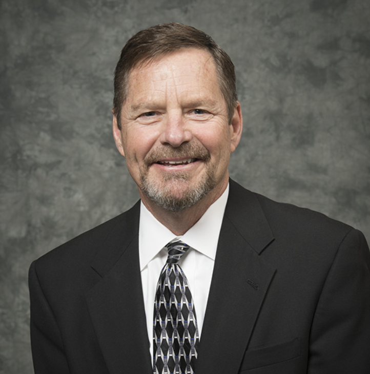 A middle-aged man with short brown hair and a trimmed beard, wearing a black suit, white shirt, and patterned tie, smiles in front of a mottled gray background.