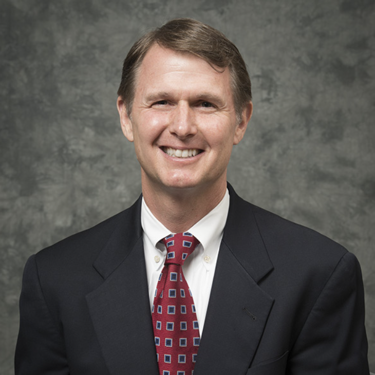 A middle-aged man with short brown hair, wearing a dark suit jacket, white dress shirt, and red patterned tie, smiles at the camera against a mottled gray background.