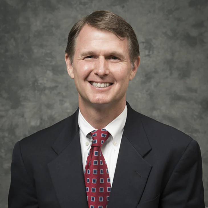 A middle-aged man with short brown hair, wearing a dark suit jacket, white dress shirt, and red patterned tie, smiles at the camera against a mottled gray background.