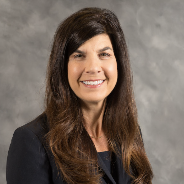 A woman with long brown hair wearing a black blazer smiles at the camera against a gray mottled background.