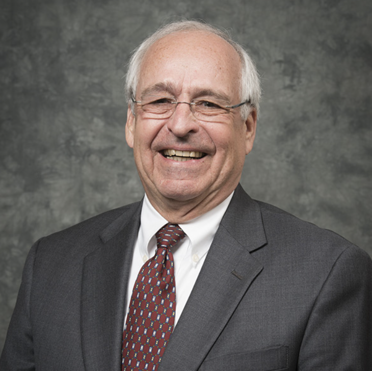 An older man with gray hair and glasses wearing a dark suit, white shirt, and patterned tie, smiling in front of a mottled gray background.