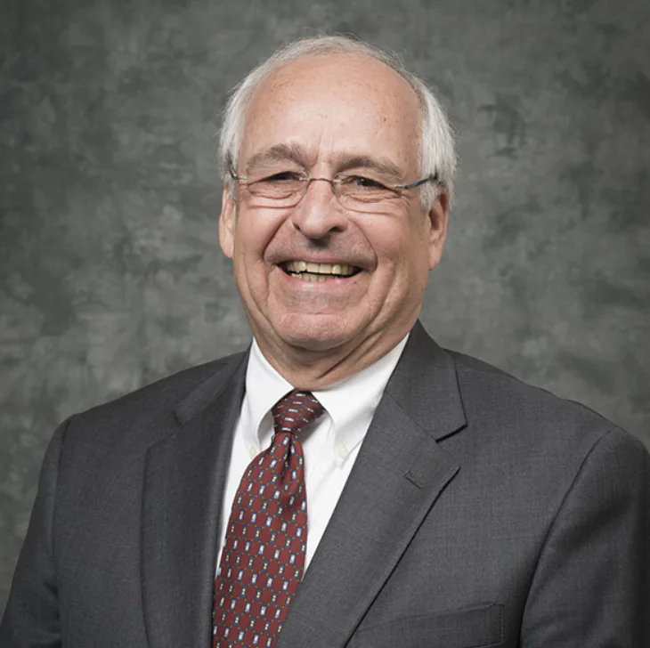 An older man with gray hair and glasses wearing a dark suit, white shirt, and patterned tie, smiling in front of a mottled gray background.