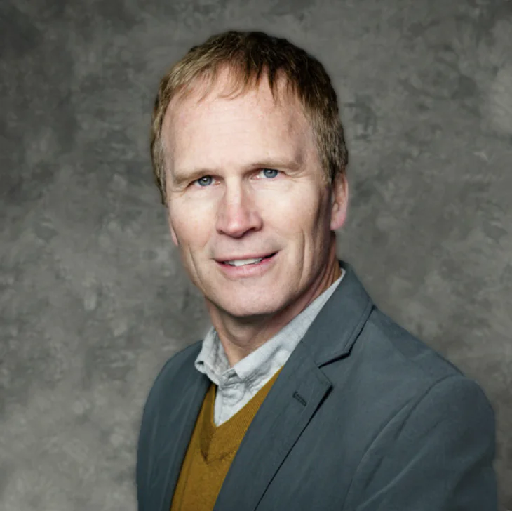 A man with short light brown hair wearing a gray blazer, mustard yellow sweater, and light-colored shirt, poses in front of a textured gray background.