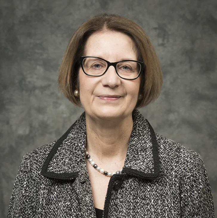 A middle-aged woman with short brown hair, wearing glasses, a gray textured jacket, a pearl necklace, and pearl earrings, stands in front of a gray mottled background, smiling slightly.