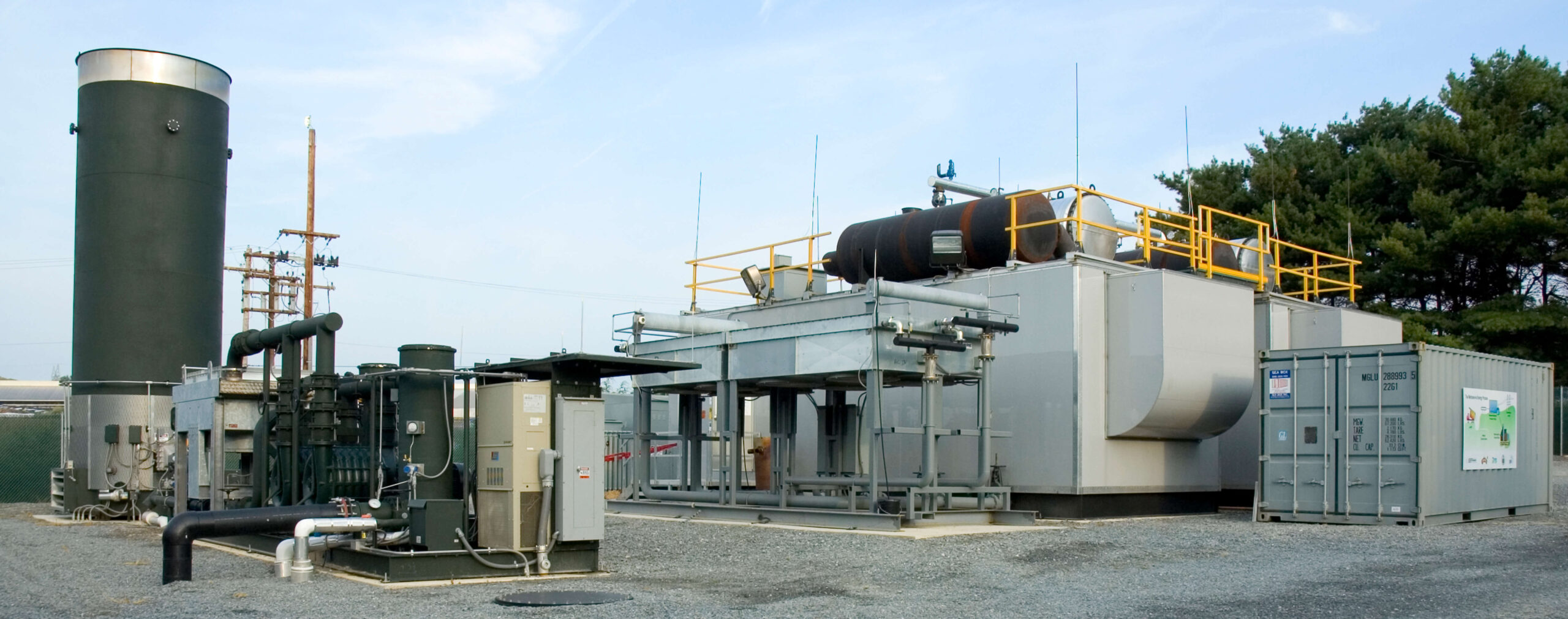 Industrial facility with metal structures, equipment, and containers on a gravel lot, surrounded by trees and utility poles under a clear sky. Yellow railings and pipes are visible on the main white building, showcasing landfill energy operations.