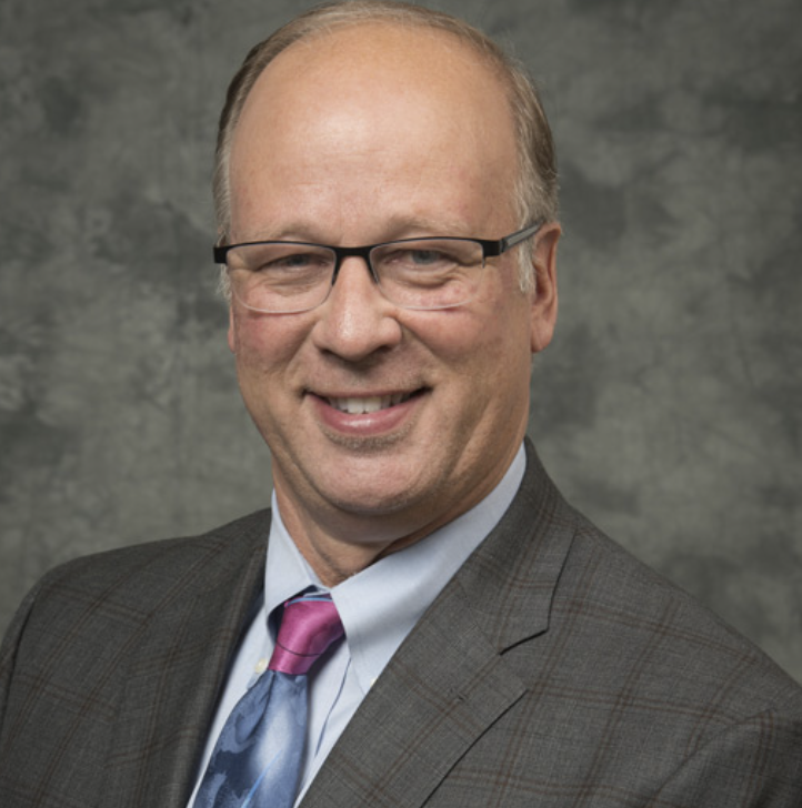 A middle-aged man with glasses, short light hair, and a plaid suit jacket smiles in front of a gray textured background. He is wearing a blue shirt and a colorful patterned tie.