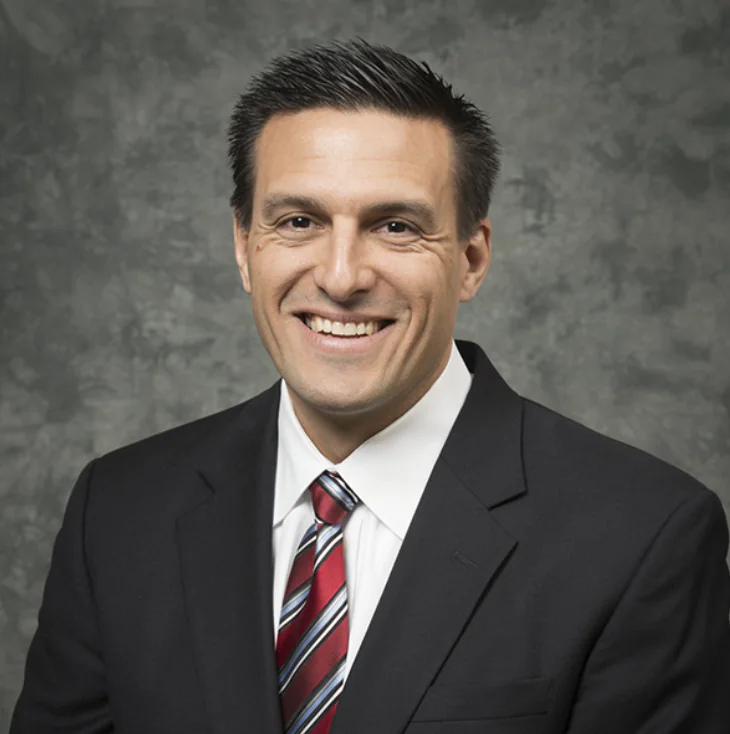 Steve Dzurik, dressed in a dark suit, white shirt, and red striped tie, smiles at the camera in front of a gray textured background.