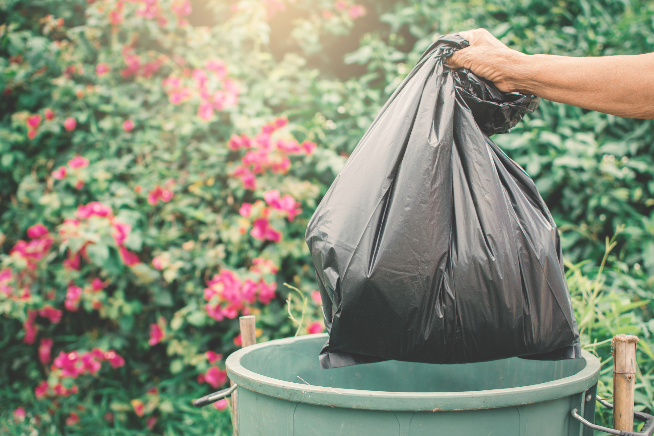 Tan hand holding plastic bag into trash. Flowers in the background