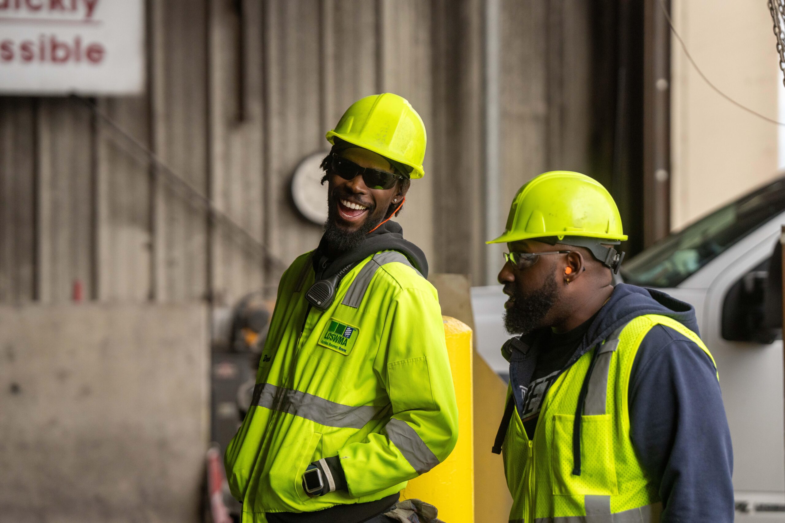 Two trash dump workers wearing neon yellow safety gear and hard hats stand together, one smiling broadly while the other looks ahead. Both wear glasses and are in an industrial setting.